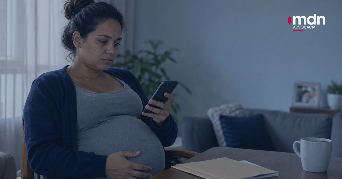 Mulher grávida pensativa com celular e documentos de demissão sobre uma mesa.